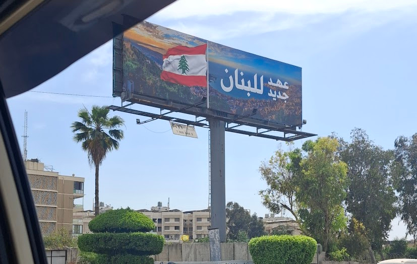 A photo taken through a car window shows a poster which displays an aerial image of the Bay of Jounieh, with text and an image of the Lebanese flag superimposed on it. The text reads: عهد جديد للبنان [translates to: A New Era for Lebanon]