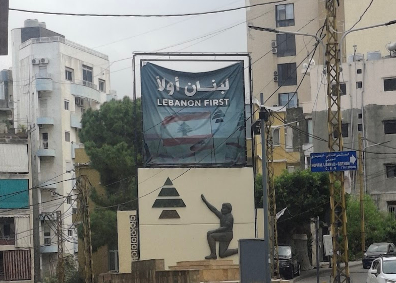 A flag showing a faded picture of the Lebanese flag and the text: 'Lebanon First', sits above a board showing a picture of a person saluting a tree