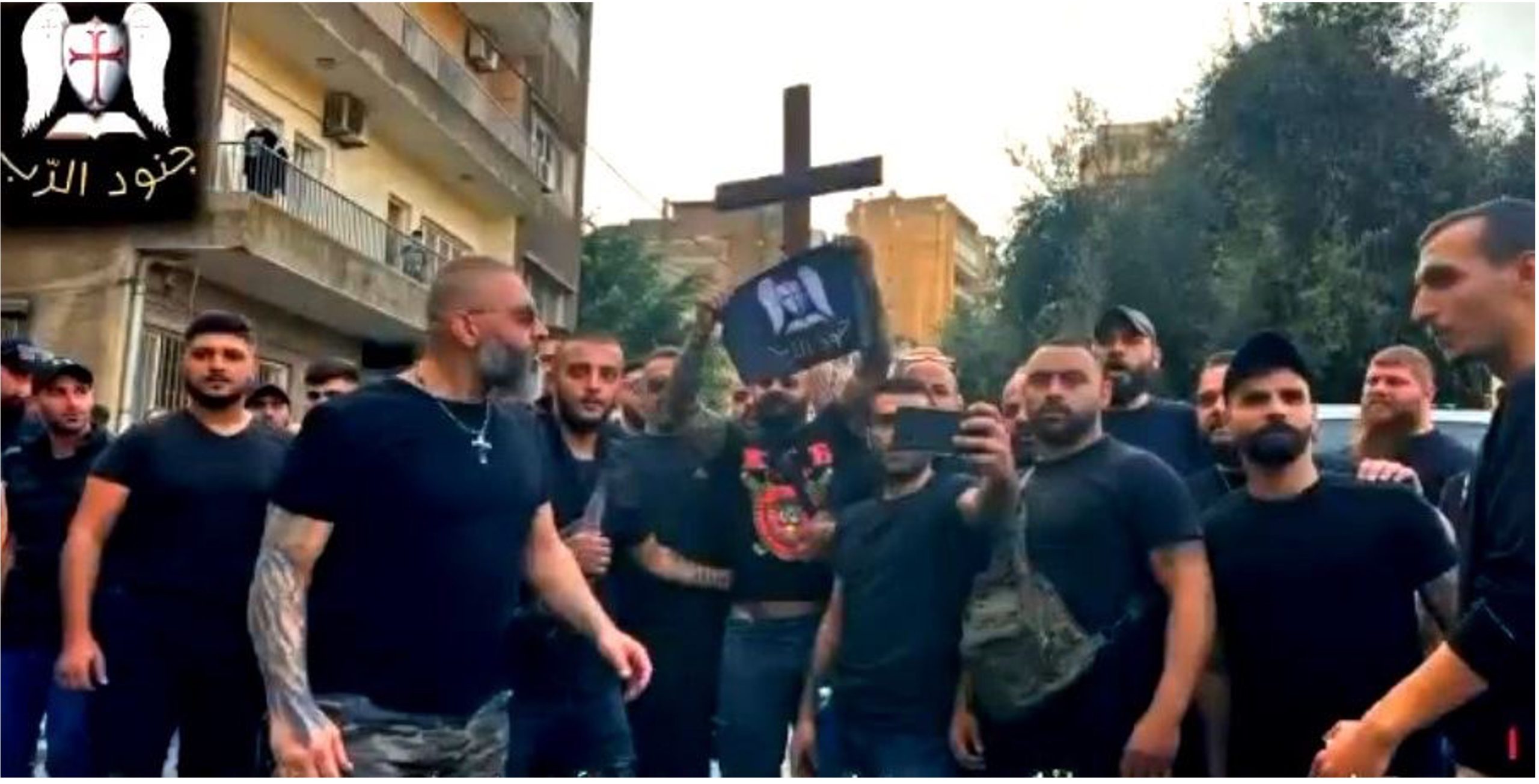 A group of men wearing black t-shirts. One is holding a flag with Soldiers of God's coat of arms of