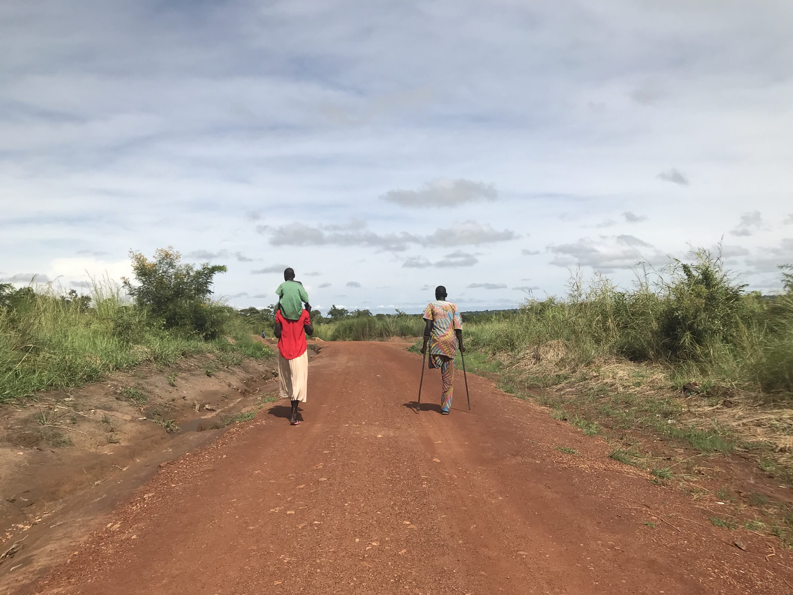 Two people walk down a dust track. One carries a young child on their shoulders. The other has an amputated leg and is using crutches.