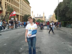 Woman in sling stands in road. People gathering behind carry Lebanese flag.